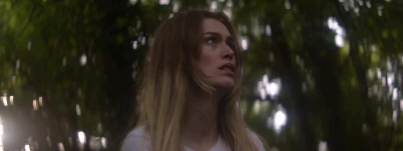 A woman turns and looks around fearfully as she stands in a grove of trees