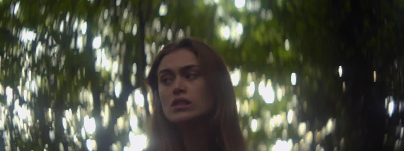 A young woman standing beneath some trees looks around with a worried expression