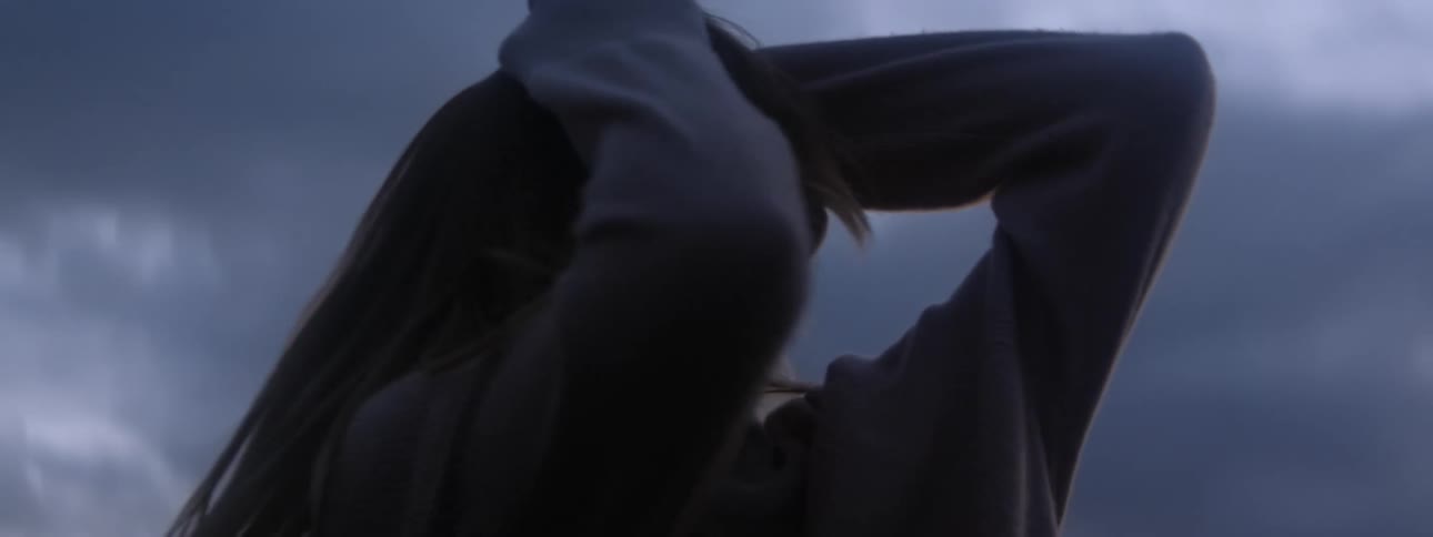 Blonde woman wearing brown sweater dances against the cloudy sky
