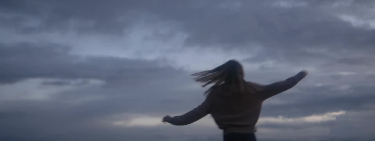 A woman in a sweater twirling outside in a field on a cloudy evening