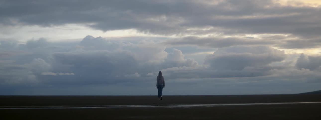 Woman with sweater and pants walks across the field against the cloudy sky at dusk