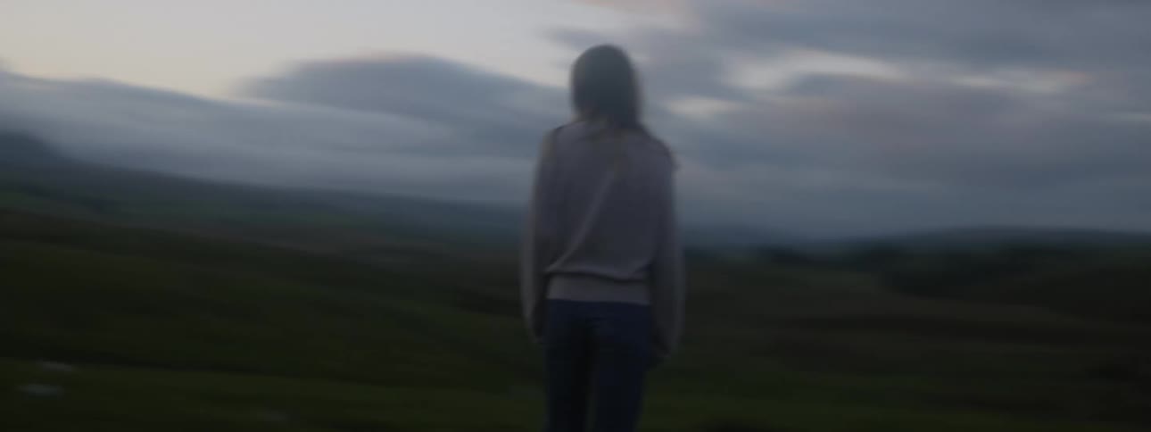 A woman stands on a hill with dark storm clouds and looks around