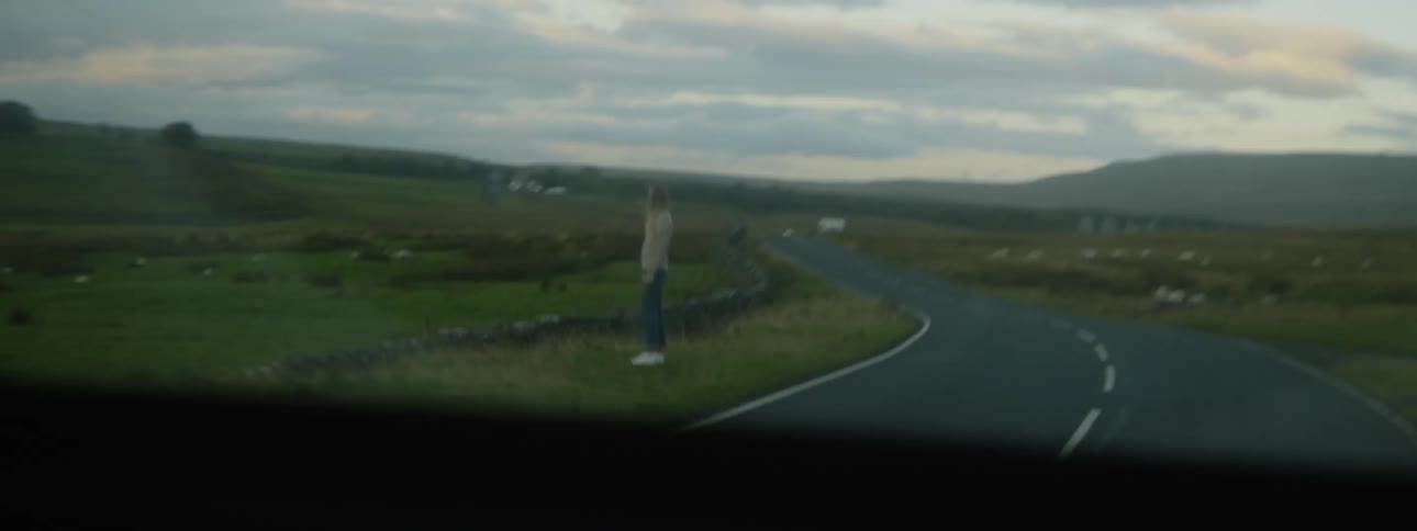 A vehicle speeds past a woman standing on the side of a rural road