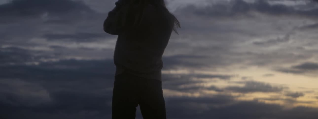Woman raises her hands to her face and turns while silhouetted against dark clouds and the red sky