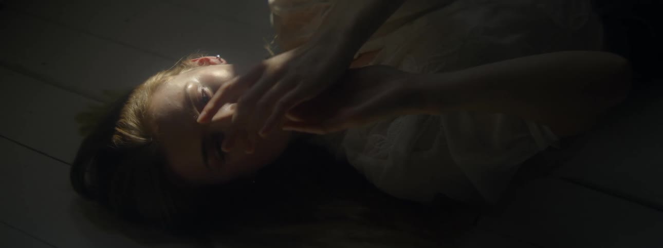 Woman lies down onfloor of shadowed room examining her hands as sun shines on her face