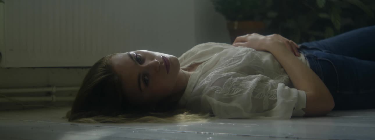 A sad looking teenager lies on a floor with her hands crossed over her stomach