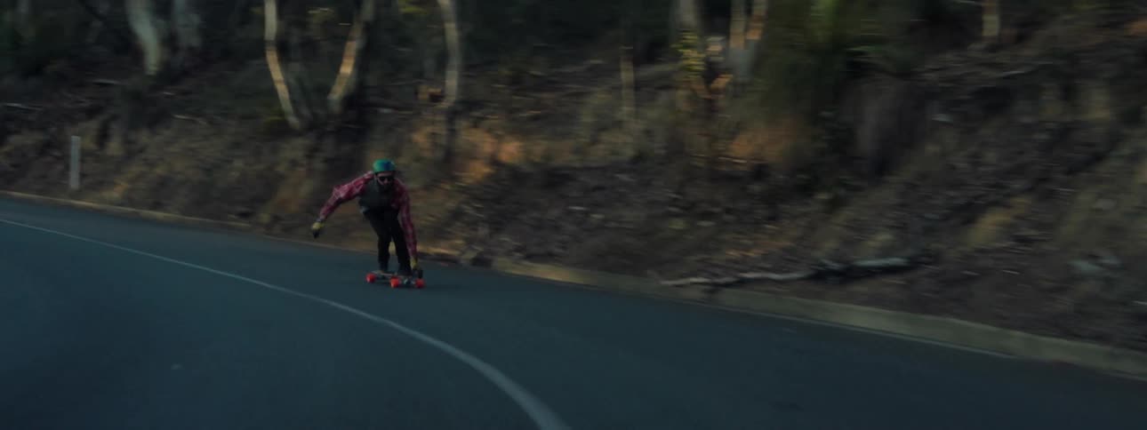 Man wearing turquoise helmet red shirt and black pants skateboards down the mountain highway