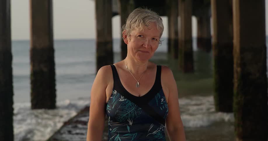 A woman wearing a bathing suit stands posing near pier pylons as ocean waves splash up