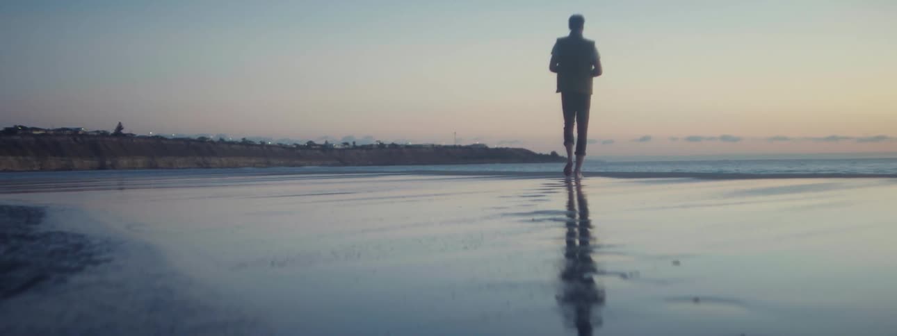A barefoot man walks along a beach in the evening