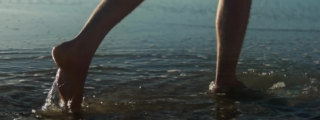 Close up of a person walking barefoot on the shallow water on the coast of a body of water at sunse