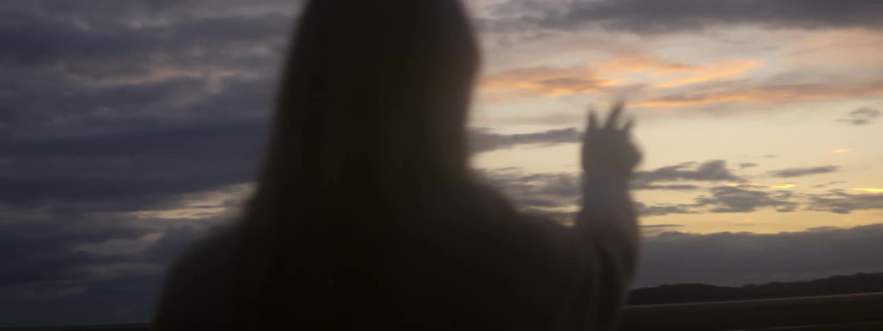 Blonde woman wearing brown sweater holds out her hand against the sky at sunset