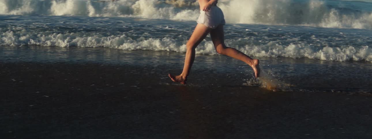 A woman is running barefoot along the beach splashing as the waves roll into shore