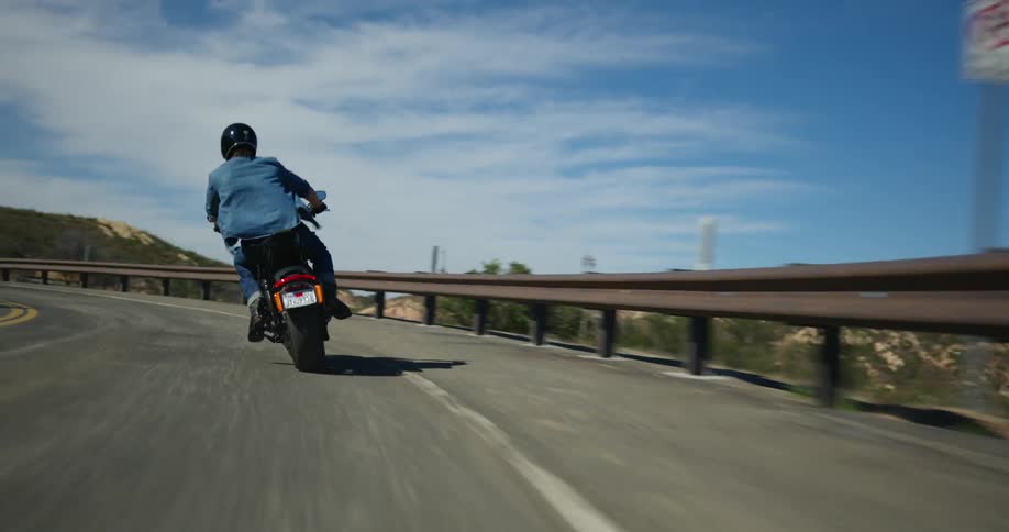 A cyclist rides a motorcycle on a road around a bend overlooking mountains in the distance