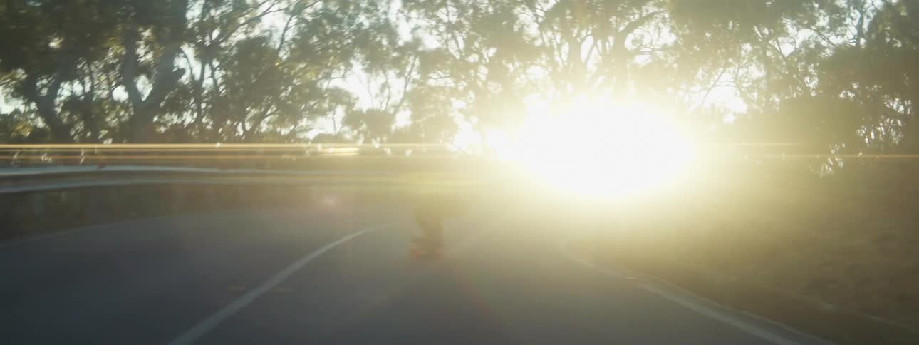 A skateboard rider is squatting down as they go around a curve and the sun is shining brightly in the distance