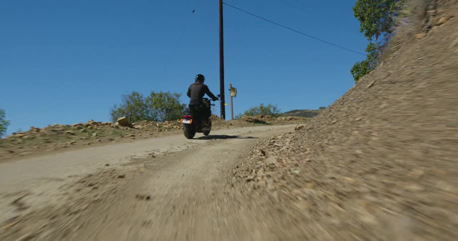 A man is shown from behind riding a motorcycle along a dirt road surrounded by mostly dirt hills and a few trees