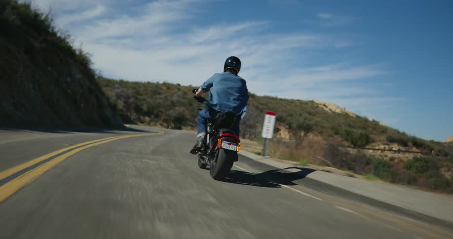 A man is shown from the back riding a motorcycle on a road surrounded by trees and grass
