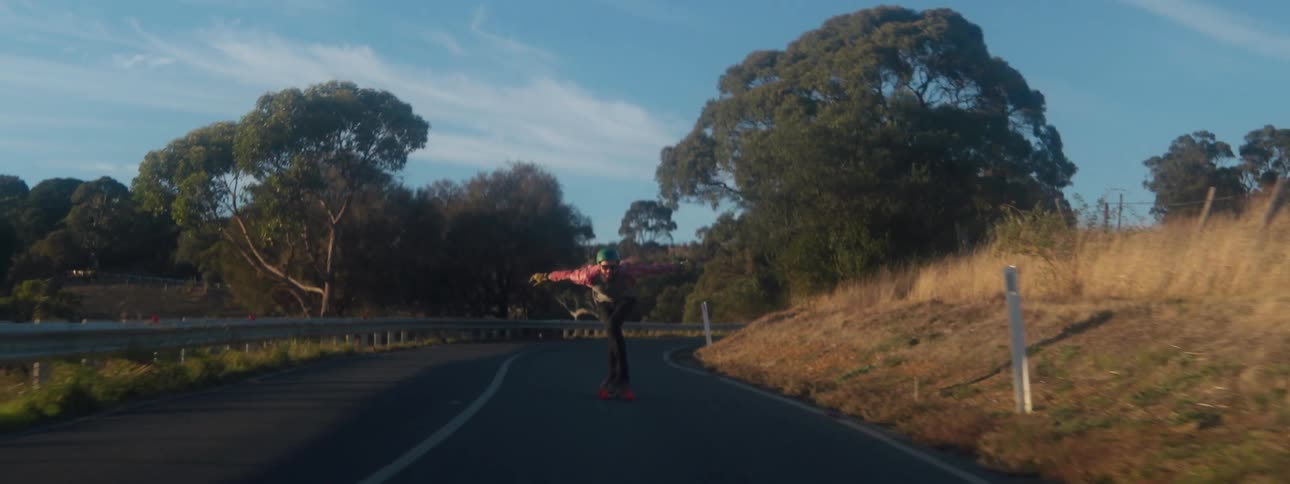 A person shown from the front skateboards down a road surrounded by trees