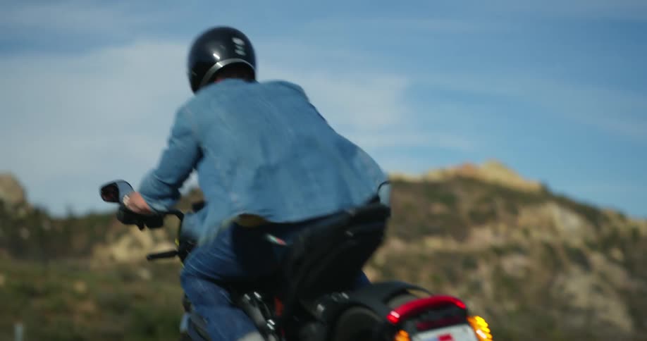 Man wearing black helmet blue shirt and jeans rides motorcycle on mountain highway