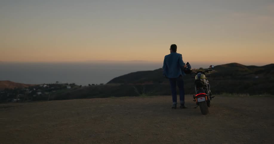 A man stands next to his parked motorcycle takes off his helmet and takes in the mountains and nature around him