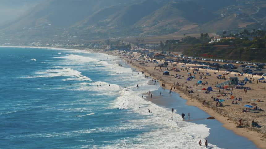 People wade into the blue ocean waters near beach bordered by parking lot and road on misty day
