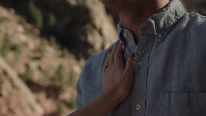 A woman slowly caresses the neck and should of her husband while they are outside
