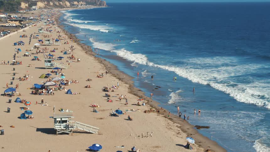 A large crowd of people line a sandy beach next to the ocean on a sunny day