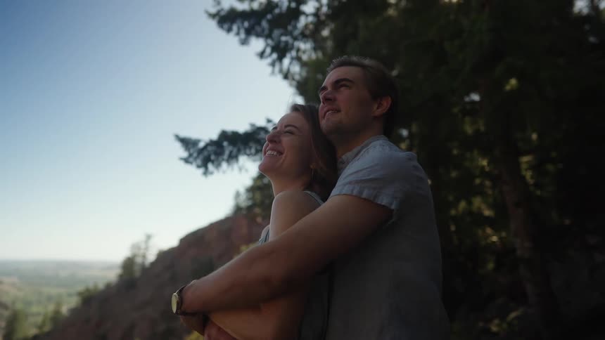 A man stands behind a woman in a forest area and hugs her as they laugh and smile together