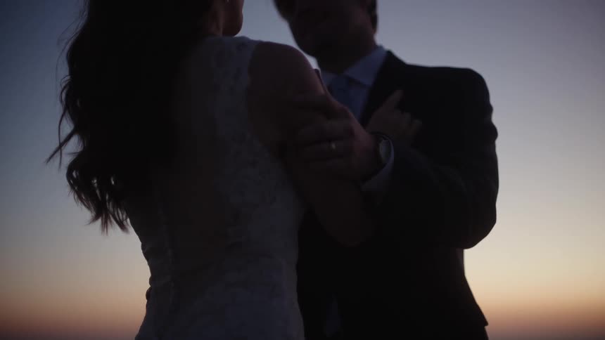 Groom wearing suit and bride in wedding dress hold each other and are silhouetted against the sky at dusk
