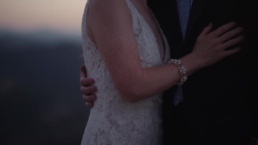 A woman wearing a lace dress and a man wearing a tuxedo stand and embrace both without their faces showing