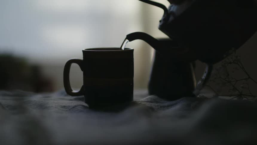 A person pours water from a kettle into a tea cup before placing the kettle on the table