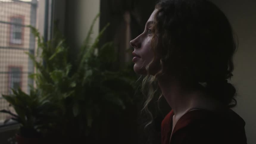 A woman with curly hair stands and looks out a window as she drinks from a mug