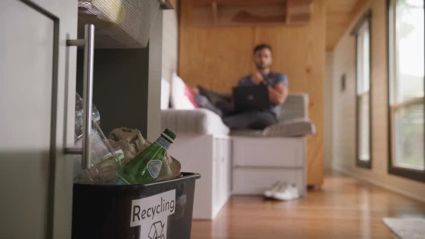 A man stares at a full recycling bin in the kitchen while working on his laptop