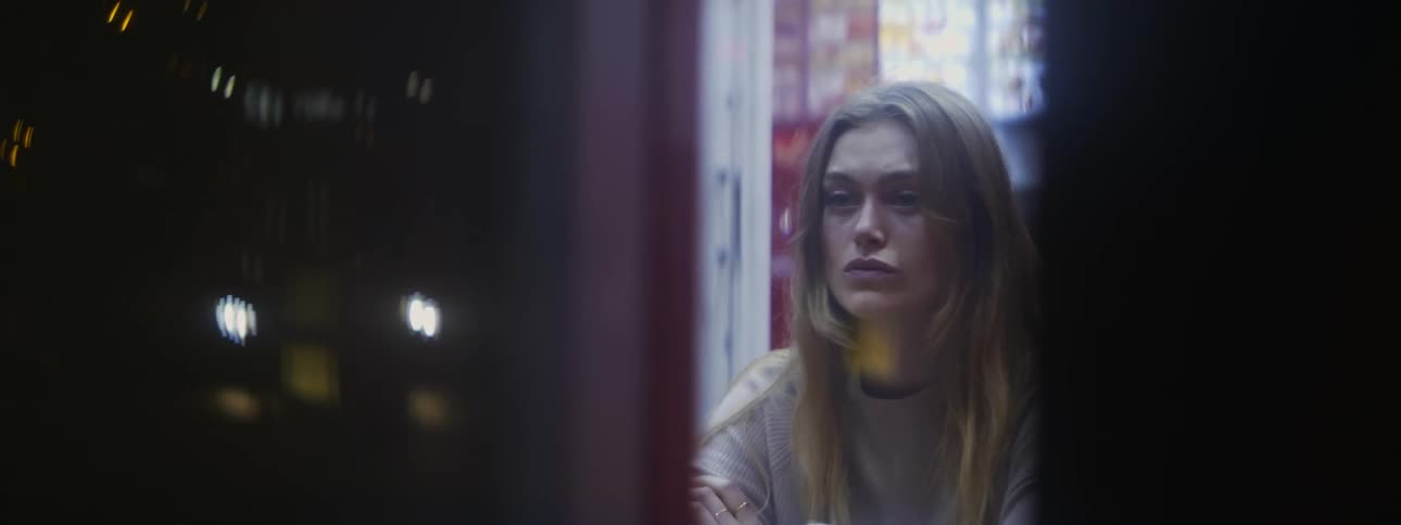 A sad looking woman sits in a cafe at night and watches a bus drive past