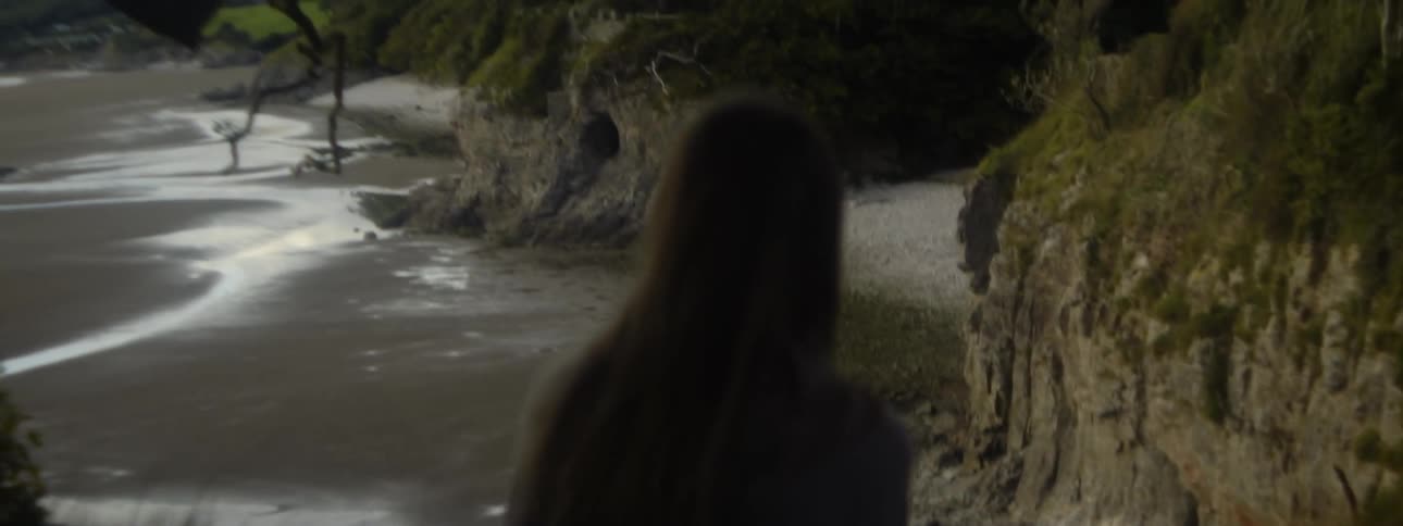 Woman sits on cliff overlooking beach with mountains in the distance