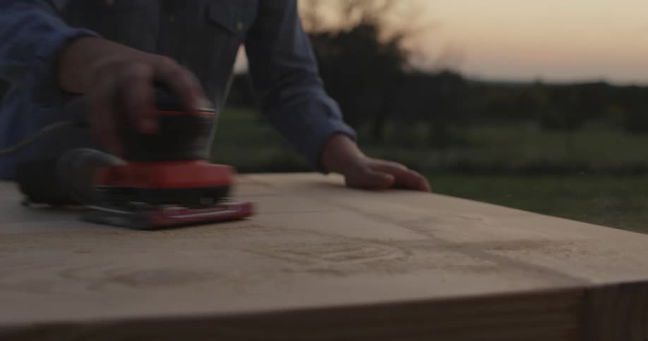 Woman stands in fenced yard in countryside sanding board with a powered sander