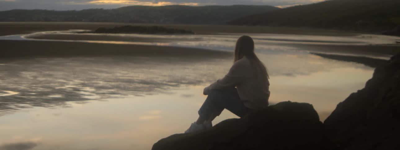 A woman sits on rocks along the shore and watches the waves