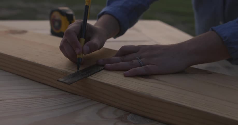 Woman uses a ruler to mark a line over a plank of wood over a workbench outdoors at evening