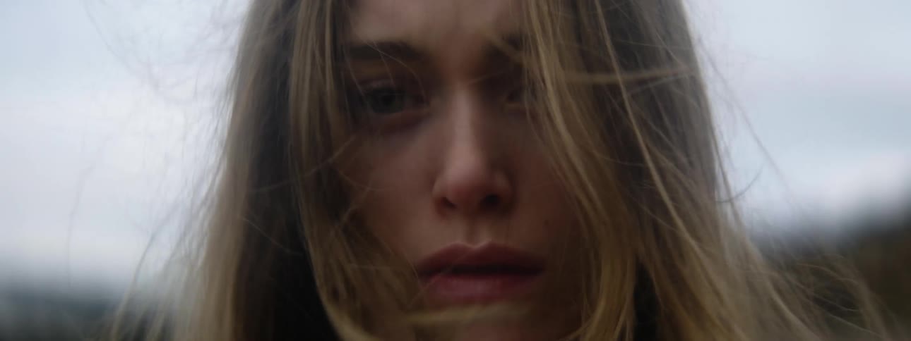 A young woman stands outdoors while looking directly into the camera with a sad look on her face