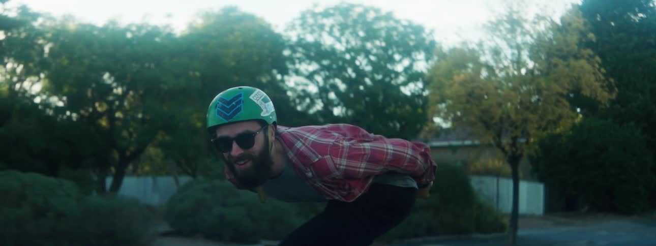 Male skateboarder wearing turquoise helmet plaid shirt and black pants skateboards down the street at dusk
