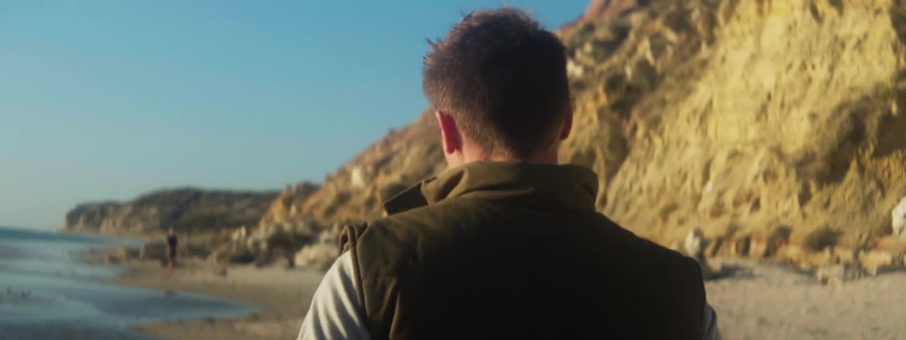 Back of a man walking on the sand beach next to the ocean close to a cliff