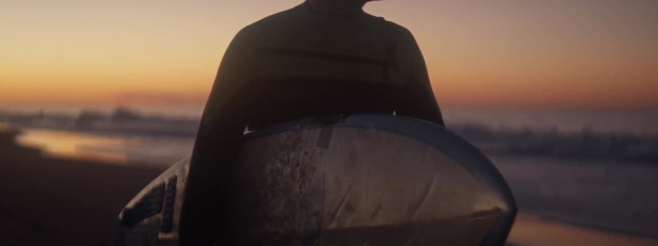 Man holding a surfboard walking along the beach during sunset