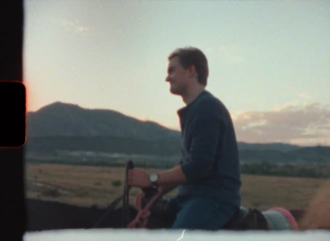Man in blue shirt jeans and woman wearing hat and white blouse sit on horse in the field at dusk in super 8 film