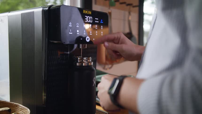 A mans arm is shown putting a thermos on a coffee making machine and pressing buttons to start it