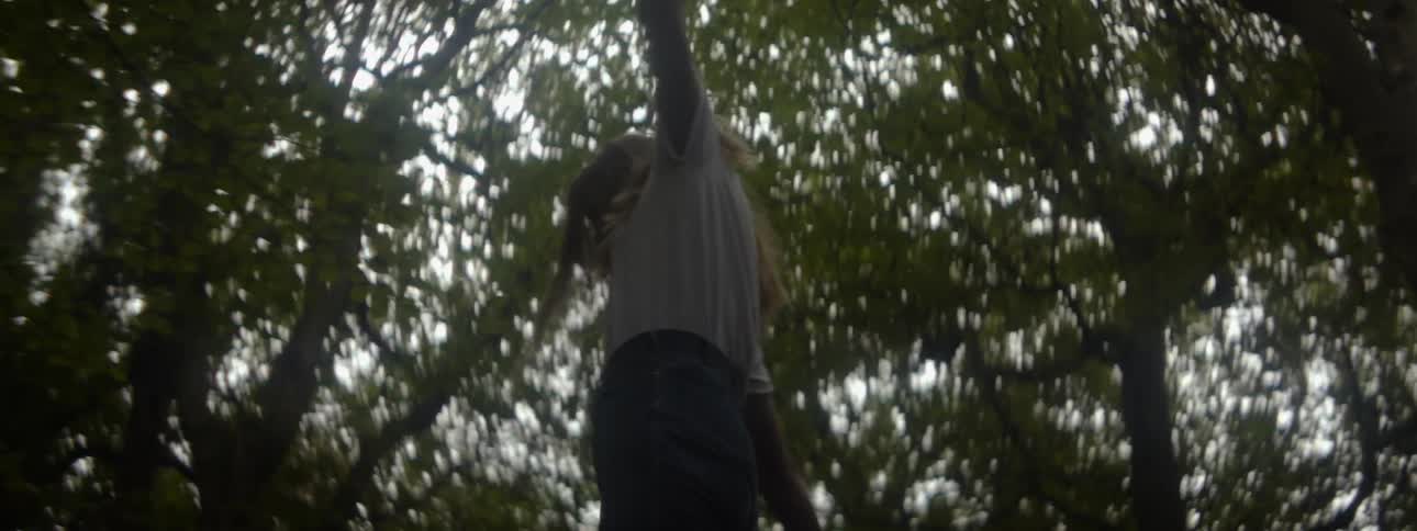 A woman stands amongst several trees and spins around carelessly with her arms stretched out to her sides