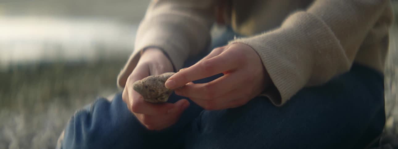 A womans hands as she sits by the water holding a rock and turning it over in her hands
