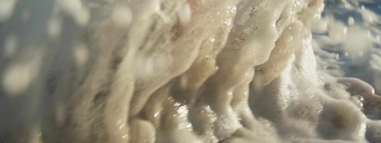 Close up of a barefoot person standing on the coast of a beach having waves crashing on the legs