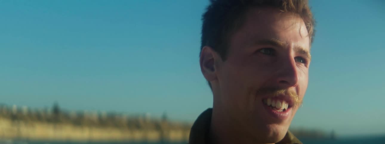 A man with a mustache walks outdoors and smiles as he looks straight ahead