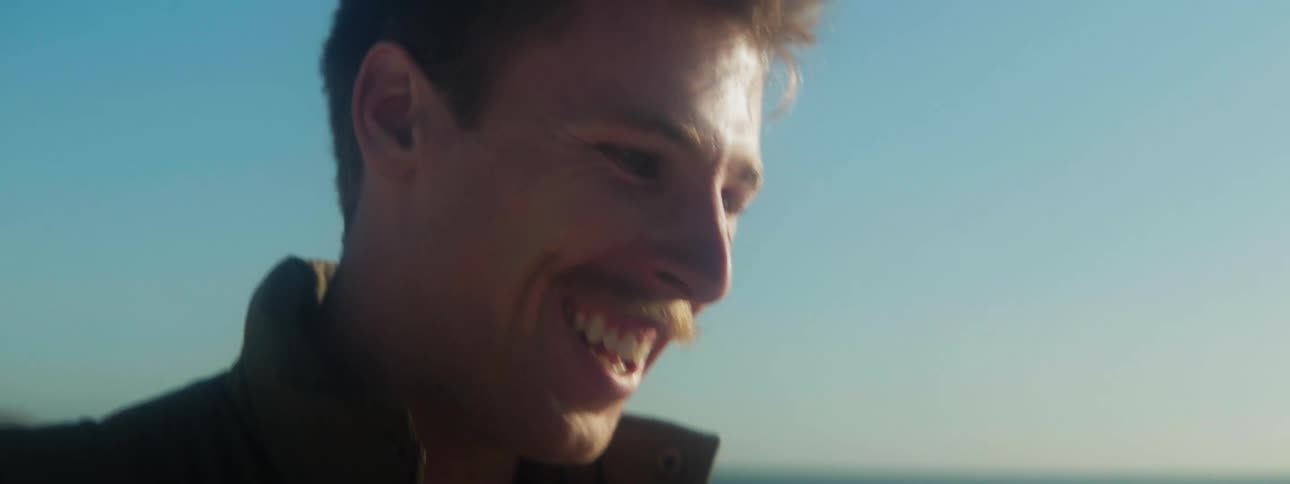 A man with a mustache smiles as he walks outside on a sunny day