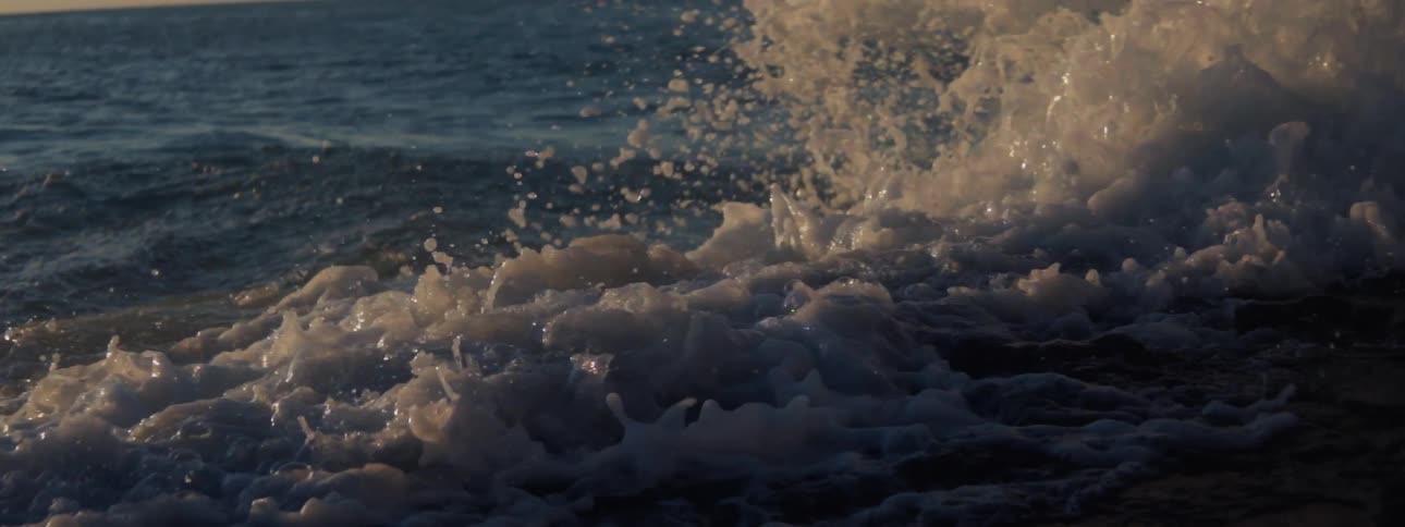 Waves crash onto rocky shore sending spray into the air and creating foam as sun shines from red tinted sky