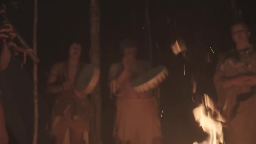 Villagers play instruments while standing around a fire at night